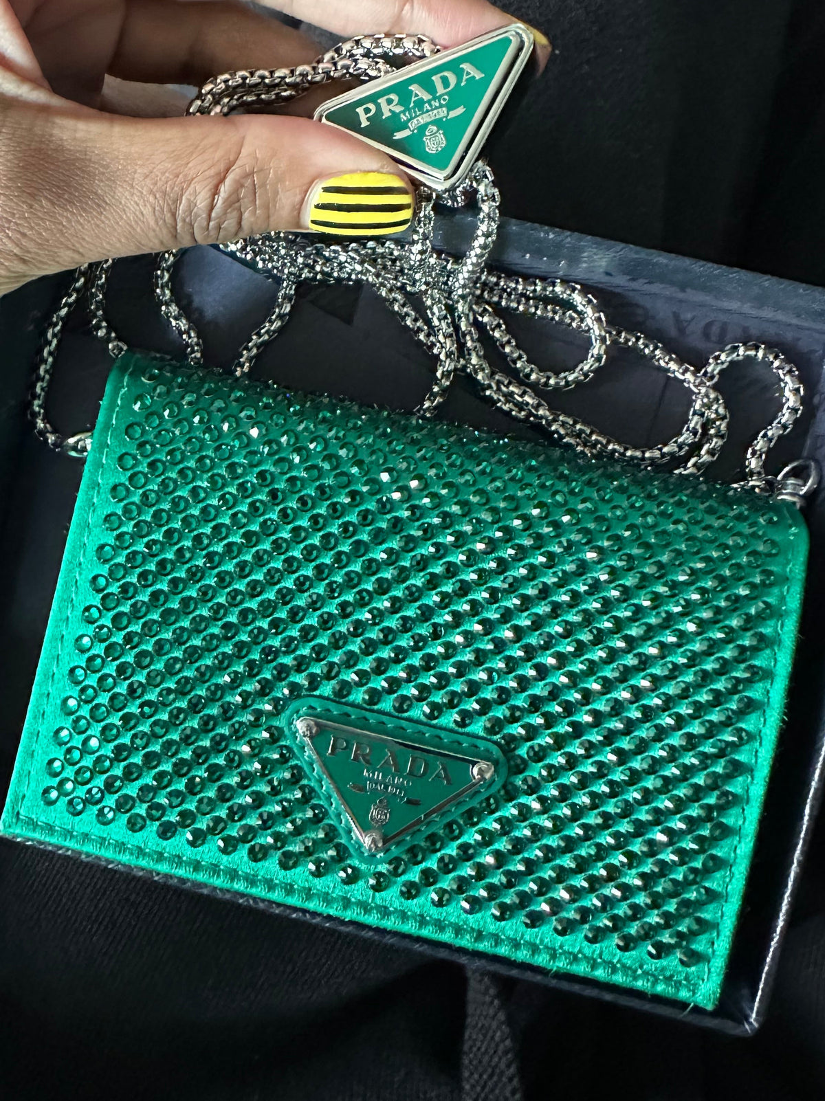 A hand holds a green Prada triangle charm with a silver chain above an open black box containing a green rhinestone-embellished Prada card holder or pouch with a matching green Prada logo plaque.